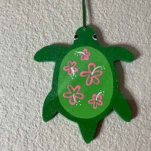 Może przedstawiać: Zielona drewniana ozdoba w kształcie żółwia z malowanym wzorem. Żółw ma zielone ciało i jest ozdobiony różowymi kwiatami hibiskusa i białymi kropkami. Do zawieszenia dołączony jest zielony sznurek.
