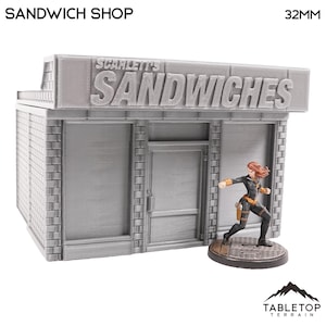Puede incluir: Una tienda de sándwiches en miniatura gris con el texto "SANDWICH SHOP" y "SCARLETT'S SANDWICHES". Una figura en miniatura de una persona con un atuendo negro está de pie frente a la tienda. La tienda está hecha de ladrillos grises.