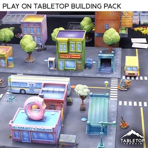 May include: A tabletop building pack with miniature buildings, trees, and vehicles. The buildings include a donut shop, a burger restaurant, and other structures. The scene is set on a grey street with crosswalks and streetlights. The text "PLAY ON TABLETOP BUILDING PACK" is at the top.