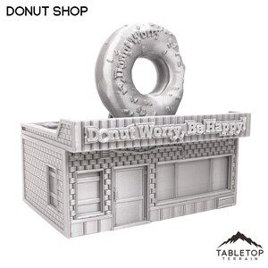 Könnte beinhalten: Graues Modell eines Donut-Shops mit einem großen Donut obenauf. Der Shop hat eine Backsteinfassade, Fenster und ein Schild mit der Aufschrift "Donut Worry, Be Happy!". Oben steht der Text "DONUT SHOP".