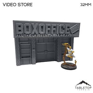 Puede incluir: Un modelo gris de una tienda de videos a escala 32 mm con un letrero que dice "BOXOFFICE ALL THE LATEST BETAMAX RELEASES". El letrero tiene un talón de boleto con la palabra "VIDEO" en él. Una figura en miniatura de un superhéroe está de pie frente a la tienda.