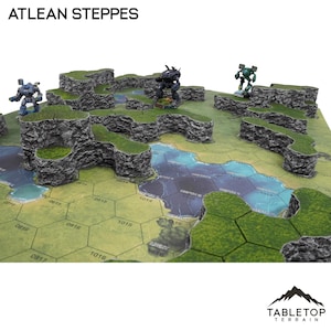 May include: A tabletop terrain set featuring rocky, elevated platforms with green grass and a blue water feature. The set includes miniature robots and the text "ATLEAN STEPPES". The terrain is designed for tabletop gaming.