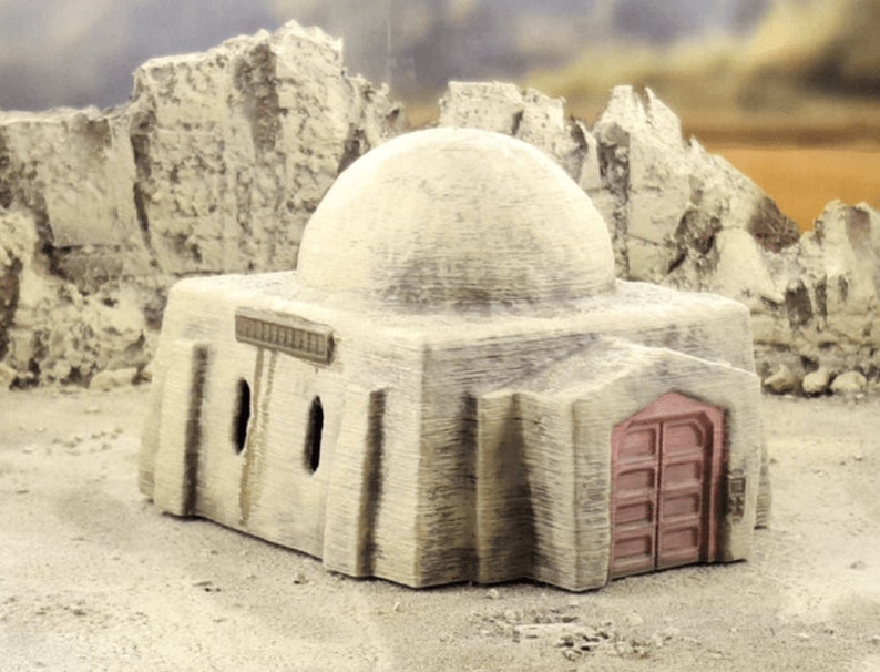 Pu&ograve; includere: Un edificio beige in miniatura con tetto a cupola e porta rossa. L'edificio ha tre piccole finestre rettangolari e un'insegna decorativa sopra le finestre. L'edificio &egrave; ambientato in un paesaggio desertico con formazioni rocciose.