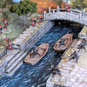Op de afbeelding: Een miniatuur oorlogsspel scène met een stenen brug over een kanaal. Rode en zwarte geverfde figuren zijn gerangschikt op de brug, de oevers van het kanaal en in twee kleine boten. De scène speelt zich af in een Japanse tuin met bomen en begroeiing.