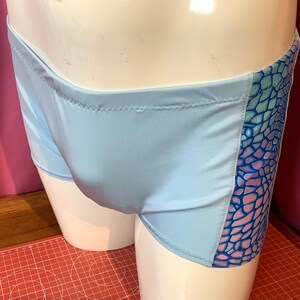Op de afbeelding: Lichtblauwe zwembroek met een zijpaneel met een schubbenpatroon in blauwe en roze tinten. De zwembroek is gemaakt van een glad, glanzend materiaal en is ontworpen om in te zwemmen.