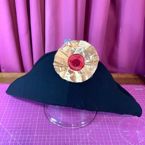 Puede incluir: Un sombrero negro con una roseta de tela dorada y roja en la parte superior. El sombrero está sobre una estera de corte púrpura.