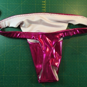 Puede incluir: Lingerie metálica rosa con forro blanco. La lencería tiene una pierna de corte alto y una cintura baja.