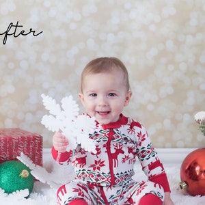 May include: A baby in a red and white Christmas pajamas holds a white snowflake ornament. The baby is sitting on a white fluffy surface with red and green Christmas ornaments and a wrapped gift box.