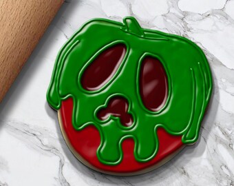 May include: A Halloween-themed cookie shaped like a poison apple, with green icing dripping over a red base. A wooden rolling pin is in the background. The cookie is on a white and gray marble surface.