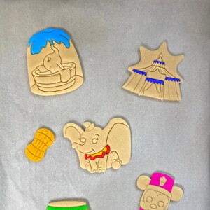 May include: A collection of Dumbo-themed cookies. The cookies include Dumbo in a bath, a circus tent, a peanut, Dumbo the elephant, a circus ring, and Mickey Mouse in a circus outfit. The cookies are tan with blue, green, yellow, and pink accents.