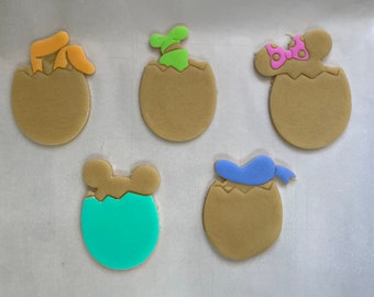 Puede incluir: Cinco galletas de azúcar con temática de Pascua. Cada galleta tiene la forma de un huevo agrietado con acentos coloridos. Las galletas presentan diseños como orejas de conejo, un brote, un lazo y un sombrero de personaje.