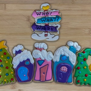 Puede incluir: Una colección de galletas de azúcar decoradas. Las galletas incluyen un poste indicador con las palabras "WHO! WHAT? WHERE!" y varias formas de casas y árboles de Navidad. Las galletas están decoradas con glaseado colorido y están dispuestas sobre una superficie de madera.