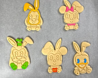 May include: Five Easter-themed biscuits shaped like cartoon characters with bunny ears. The biscuits feature characters with colourful accents, including yellow, pink, green, and orange details. The biscuits are arranged on a light-coloured surface.