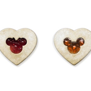May include: Two heart-shaped cookies with a Mickey Mouse head cutout filled with red and orange jelly. The cookies have a light-colored, glittery exterior and are set against a white background.