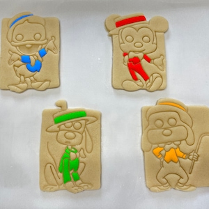 May include: Four unbaked cookies with cartoon character designs. The cookies feature Donald Duck with a blue scarf, Mickey Mouse in red attire, a dog in a green outfit, and a dog with a yellow vest and cane. The cookies are rectangular.