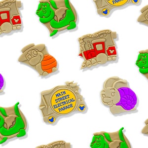 May include: Assortment of tan-colored, cartoon-style character-shaped cookies. Designs include a train, a dragon, a snail, and a turtle. One cookie features the text "Main Street Electrical Parade". Each cookie has colorful accents.