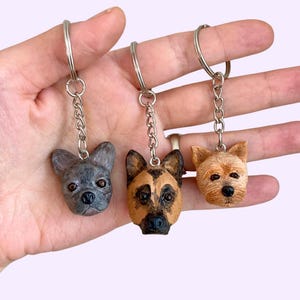 May include: Three dog head keyrings. A grey French Bulldog, a brown and black German Shepherd, and a light brown Yorkshire Terrier.