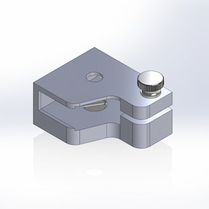 May include: A silver metal clamp with a small knob on the top. The clamp has a rectangular shape with a cut-out section on one side.