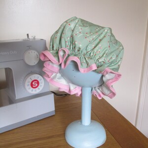May include: A mint green shower cap with a floral pattern and pink trim, displayed on a light blue stand. The cap is ruffled and sits atop a round base and a vertical post. A sewing machine is visible in the background.