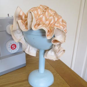 May include: A vintage bonnet in peach with a white floral design, displayed on a light blue stand. The bonnet has a cream-colored, ruffled edge. The stand has a round base and a long stem. The bonnet is likely made of cotton.