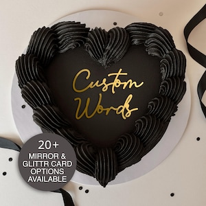 May include: A heart-shaped cake with black frosting and the words "Custom Words" in gold script. The cake is surrounded by black frosting swirls and sits on a white plate. A circular graphic states "20+ Mirror & Glitter Card Options Available."