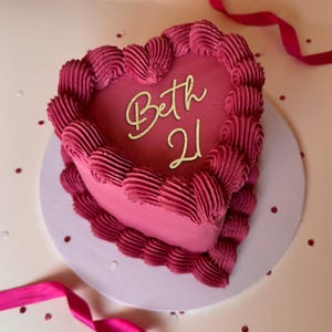 May include: A heart-shaped cake decorated with pink frosting and a gold inscription that reads "Beth 21".
