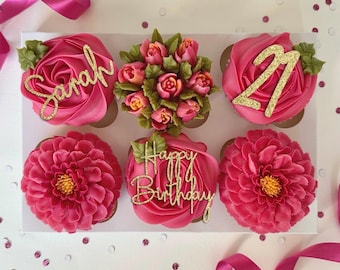 May include: A box of six pink cupcakes decorated with gold glitter lettering. The cupcakes are decorated with a variety of designs, including flowers, the name "Sarah", and the phrase "Happy Birthday".