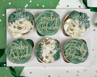 May include: Six green and white cupcakes with gold glitter and the words "Baby Johnson" on top. The cupcakes are arranged in a white box.