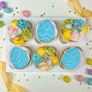 May include: Six Easter cupcakes in a white box, each with pastel frosting and decorative elements. Two cupcakes feature blue egg designs, while others have floral and sprinkle toppings. Yellow and white checkered ribbon and pastel eggs surround the box.