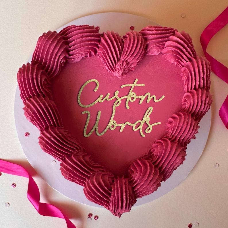 May include: A heart-shaped cake with pink frosting and a gold glitter inscription that reads "Custom Words".
