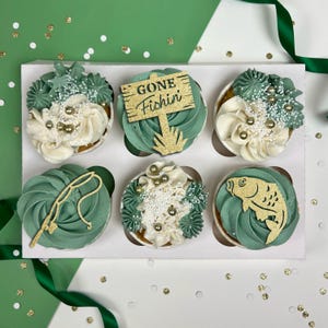 May include: Six green and white cupcakes decorated with fishing-themed toppers. The cupcakes are arranged in a white cupcake holder. The toppers include a fishing rod, a fish, and a sign that says "Gone Fishin'."