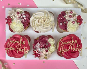 Può includere: Sei cupcake decorati con glassa rossa e bianca, codette dorate e lettere dorate commestibili. I cupcake sono disposti in un portacupcake bianco. I cupcake sono decorati con le parole "Sarah's Hen Party".