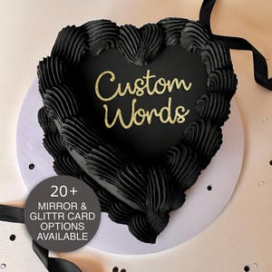 May include: A heart-shaped cake with black frosting and the words "Custom Words" in gold glitter. The cake is on a white plate, with black ribbon and a circular graphic that says "20+ Mirror & Glitter Card Options Available."