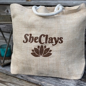 May include: A beige tote bag with a white rope handle and a brown logo that says "SheClays" with a brown lotus flower design.