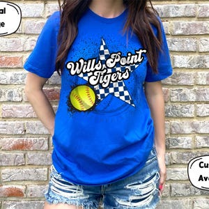 Puede incluir: Una camiseta azul real con un diseño de estrella a cuadros blancos y negros. La estrella tiene el texto "Wills Point Tigers" en un contorno blanco. Una pelota de softball amarilla es parcialmente visible detrás de la estrella.