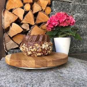 Puede incluir: Un pastel glaseado de chocolate con rodajas de almendras en una tabla de madera. A la derecha, una hortensia rosa en una maceta blanca, con una pila de leña al fondo. El pastel es el punto focal.