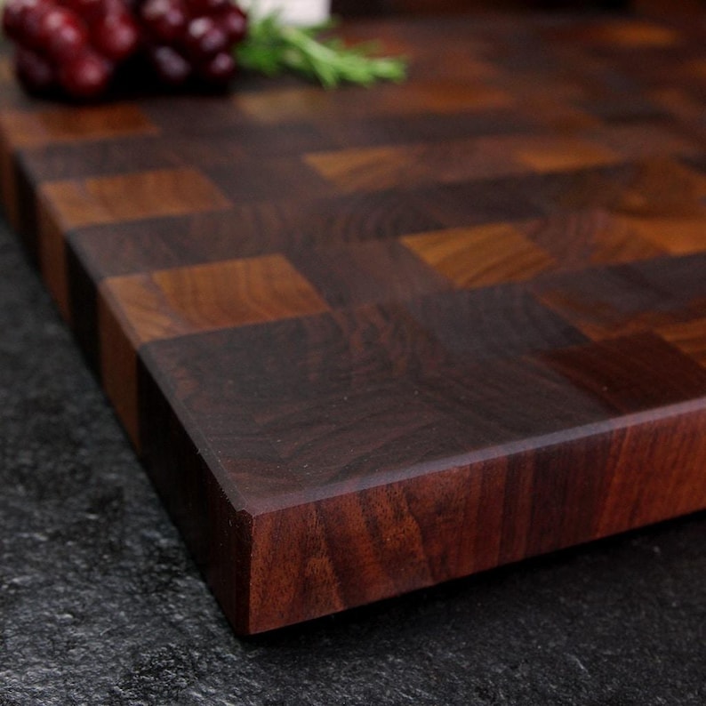 May include: A rectangular wooden cutting board featuring an end-grain construction with a dark brown checkerboard pattern. The surface displays visible wood grain textures and natural variations in colour, with a cluster of red grapes in the corner.