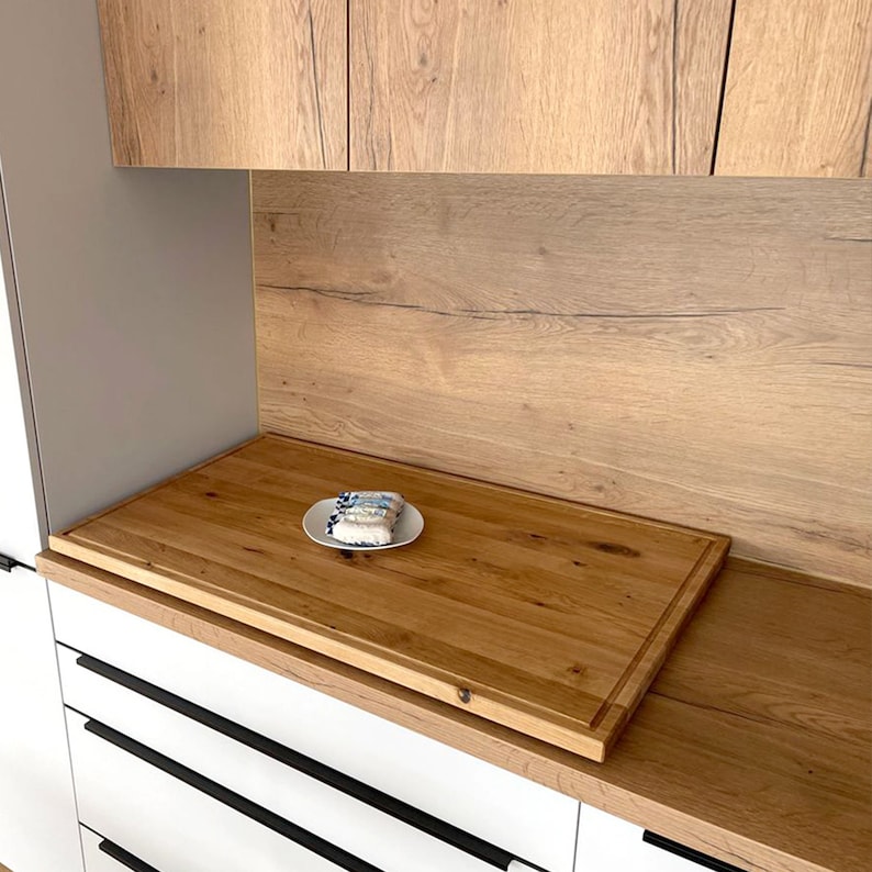 May include: A large, rectangular wooden cutting board with a light brown finish sits on a white countertop. The board has a slightly raised edge and is made of multiple pieces of wood.