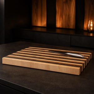 Peut inclure: Une planche &agrave; d&eacute;couper rectangulaire en bois pr&eacute;sentant des rayures horizontales altern&eacute;es de tons bois clair, moyen et fonc&eacute;. Un couteau de cuisine en acier inoxydable avec un manche argent&eacute; repose en diagonale sur la surface.