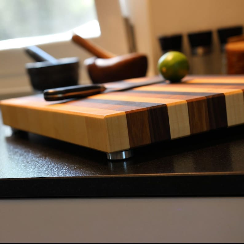 Peut inclure: Une planche &agrave; d&eacute;couper rectangulaire en bois compos&eacute;e de bandes altern&eacute;es de bois clair et fonc&eacute;. La planche est sur&eacute;lev&eacute;e sur de petits pieds en m&eacute;tal argent&eacute; et supporte un couteau de cuisine ainsi qu'un citron vert pos&eacute; sur sa surface.