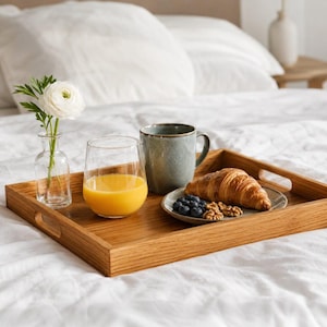 May include: A rectangular wooden serving tray holds a clear glass of orange juice, a speckled grey ceramic mug, and a small plate with a golden-brown croissant, fresh blueberries, and walnuts, alongside a small glass vase with a white flower.