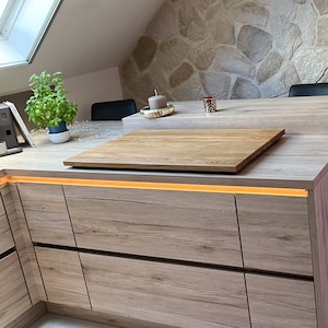May include: A wooden cutting board sits on top of a light brown countertop with a strip of LED lights underneath. The countertop is part of a kitchen island with drawers.