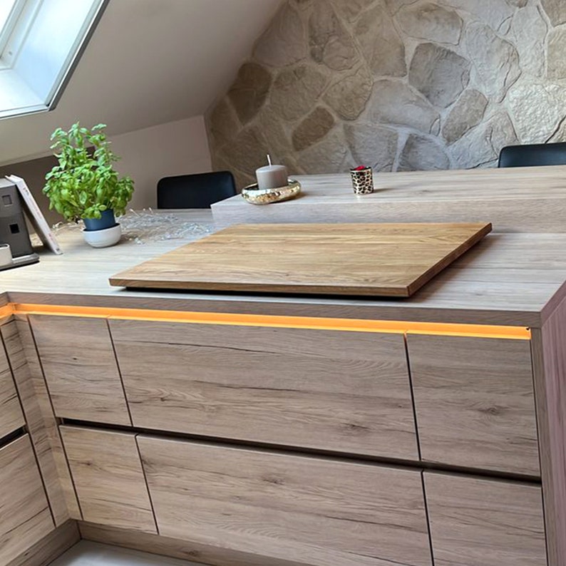 May include: A wooden cutting board sits on top of a light brown countertop with a strip of LED lights underneath. The countertop is part of a kitchen island with drawers.