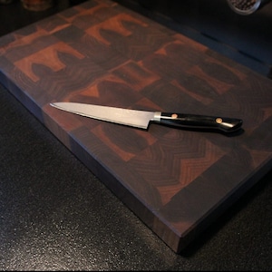 May include: A rectangular end-grain wooden chopping board featuring a dark and medium brown checkerboard pattern. A stainless steel kitchen knife with a black handle and two copper-coloured rivets rests diagonally across the board surface.