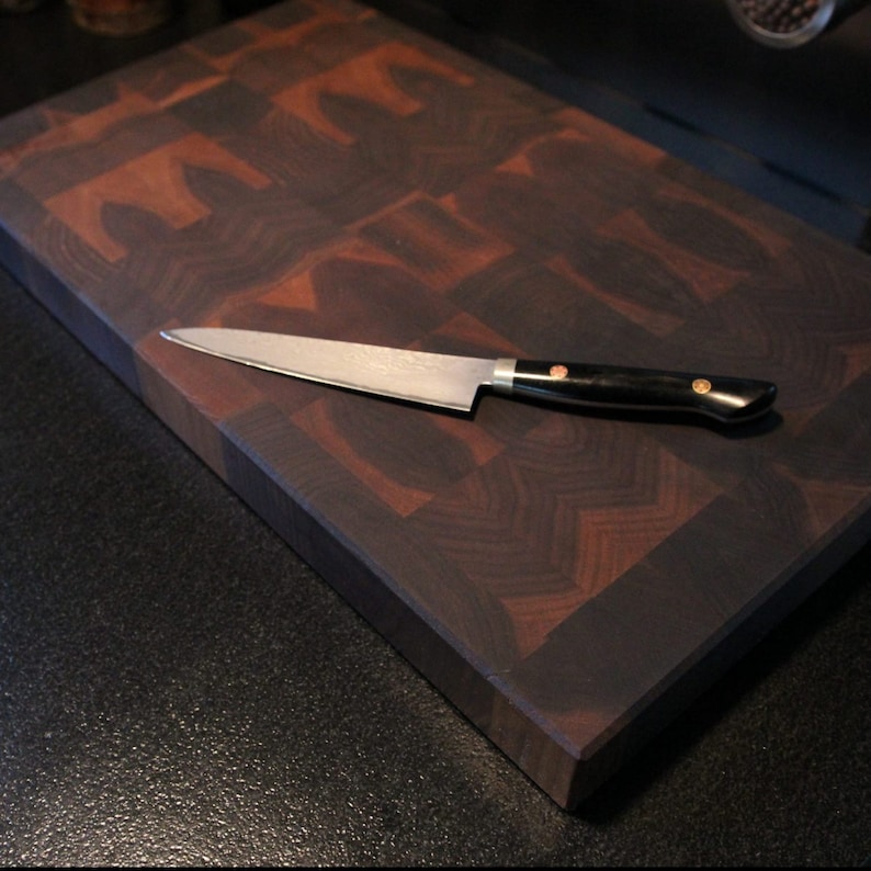 May include: A rectangular end-grain wooden chopping board featuring a dark and medium brown checkerboard pattern. A stainless steel kitchen knife with a black handle and two copper-coloured rivets rests diagonally across the board surface.