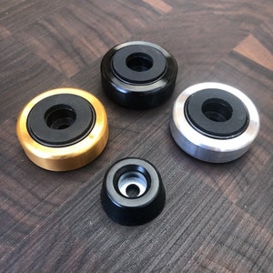 May include: Four circular components consisting of three larger metal rings in gold, black, and silver finishes, each featuring a central black rubber insert, and one smaller, tapered black metal component with a central hole and silver interior.