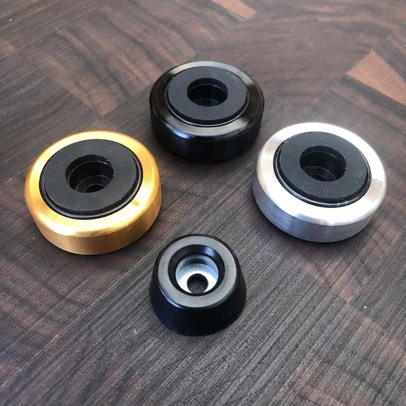 May include: Four circular components consisting of three larger metal rings in gold, black, and silver finishes, each featuring a central black rubber insert, and one smaller, tapered black metal component with a central hole and silver interior.