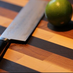 Peut inclure: Une planche &agrave; d&eacute;couper rectangulaire pr&eacute;sentant des bandes horizontales altern&eacute;es de bois clair, moyen et brun fonc&eacute;. Un couteau de cuisine en m&eacute;tal avec un manche noir repose en diagonale sur la planche, &agrave; c&ocirc;t&eacute; d'un citron vert entier.