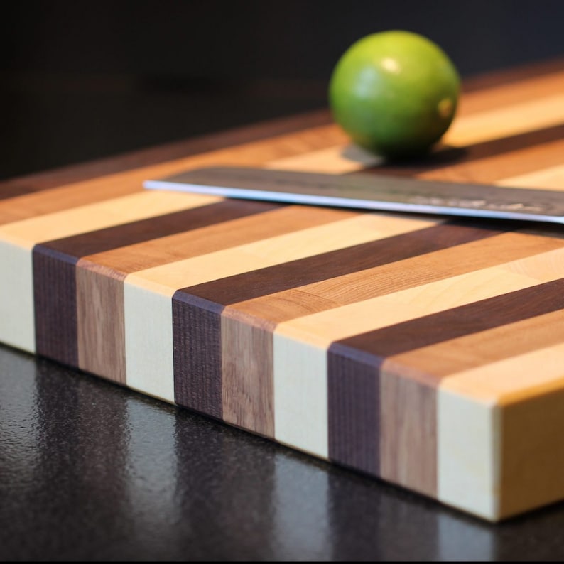 Peut inclure: Une planche &agrave; d&eacute;couper rectangulaire en bois pr&eacute;sentant des bandes altern&eacute;es de bois cr&egrave;me clair, brun moyen et brun fonc&eacute;. Une lame de couteau en m&eacute;tal argent&eacute; repose horizontalement sur la surface et un citron vert est pos&eacute; pr&egrave;s du bord arri&egrave;re.