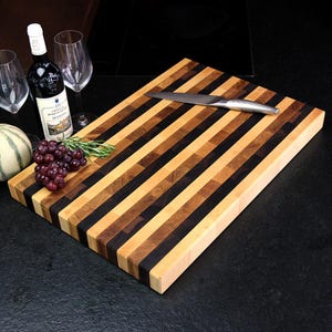 Peut inclure: Une planche &agrave; d&eacute;couper rectangulaire en bois pr&eacute;sentant des bandes altern&eacute;es de grain de bois clair, moyen et brun fonc&eacute;. Un couteau de cuisine en m&eacute;tal argent&eacute; repose sur la surface. Une bouteille de vin rouge, deux verres et du raisin.